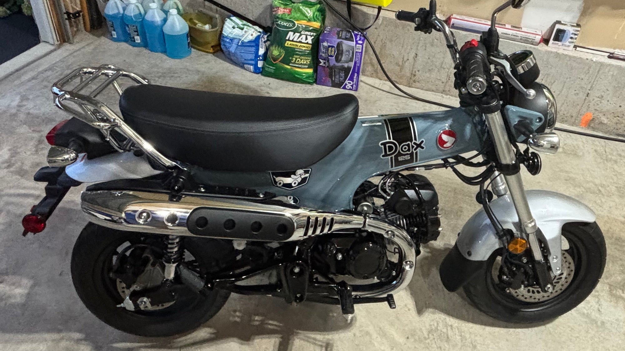 A Honda DAX 125, parked in a garage. The bike features the OEM Honda Pearl Gray finish, a plain black seat, chrome exhaust and mirrors, and a chrome rear luggage rack.